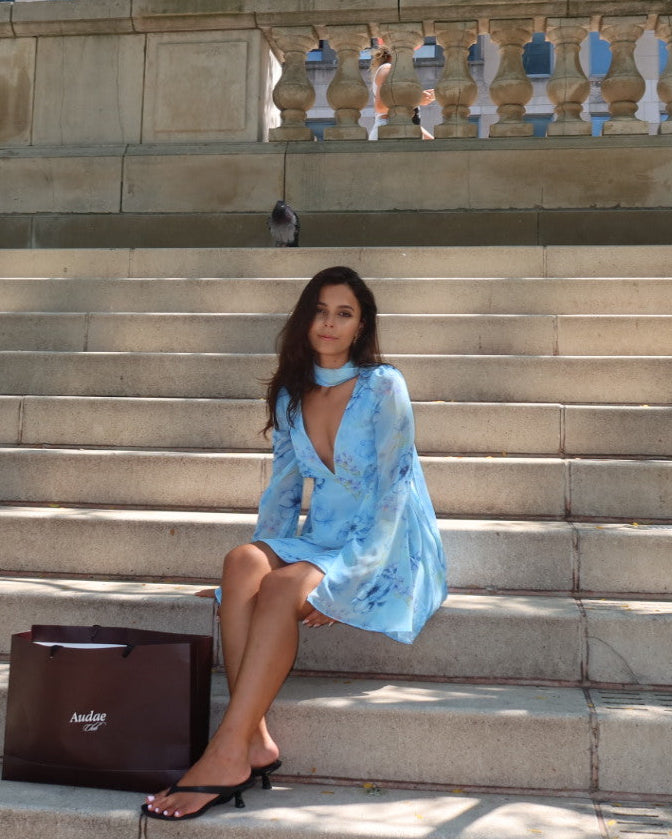 Woman in a light blue dress sitting on stone steps with a tree and city buildings in the background.