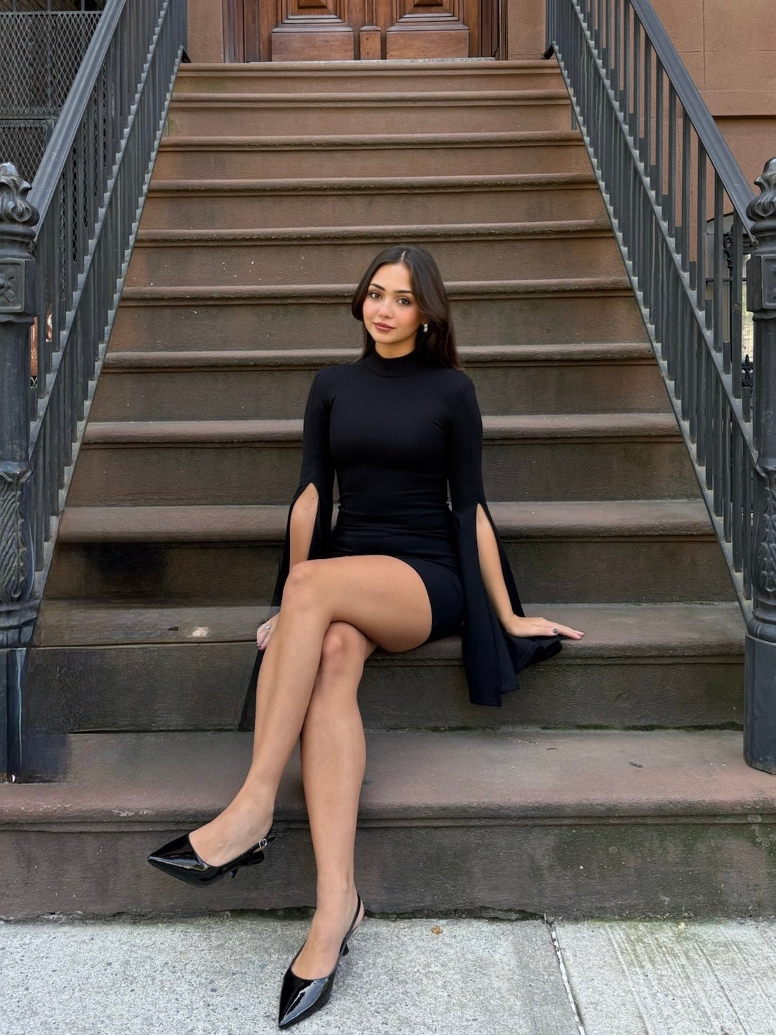 Woman in a black dress sitting on steps in front of a brownstone building.