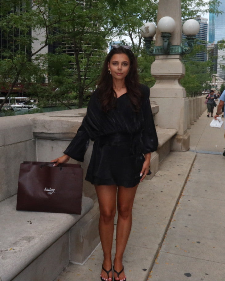 Woman in black outfit holding a brown bag on a city sidewalk with trees and buildings in the background.