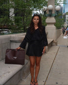 Woman in black outfit holding a brown bag on a city sidewalk with trees and buildings in the background.