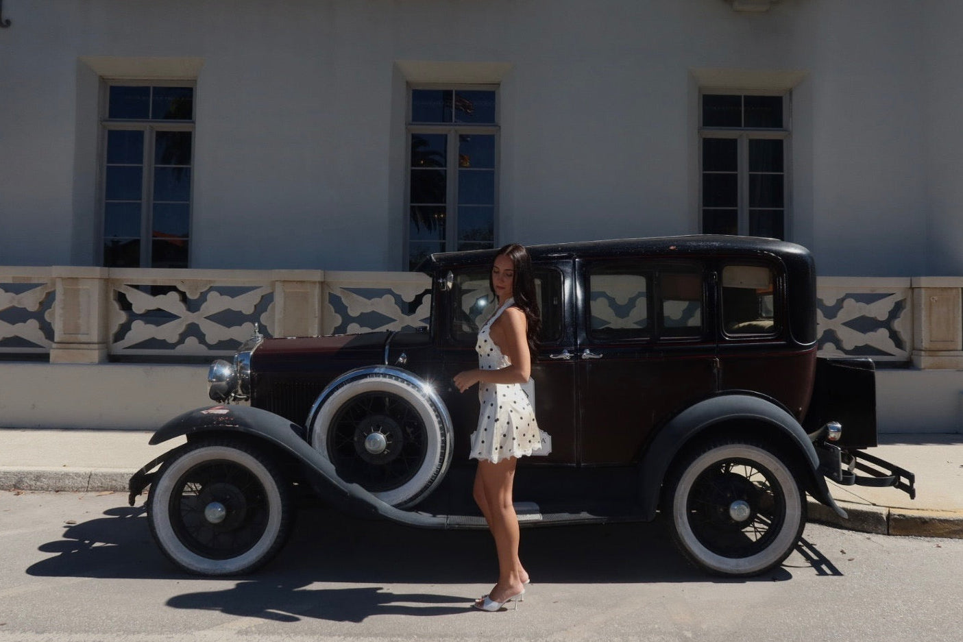 Woman standing next to an old-fashioned car in front of a building with decorative elements.