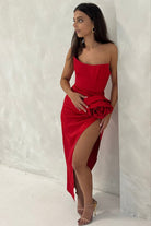Woman in a red one-shoulder dress with a high slit, standing against a white wall.