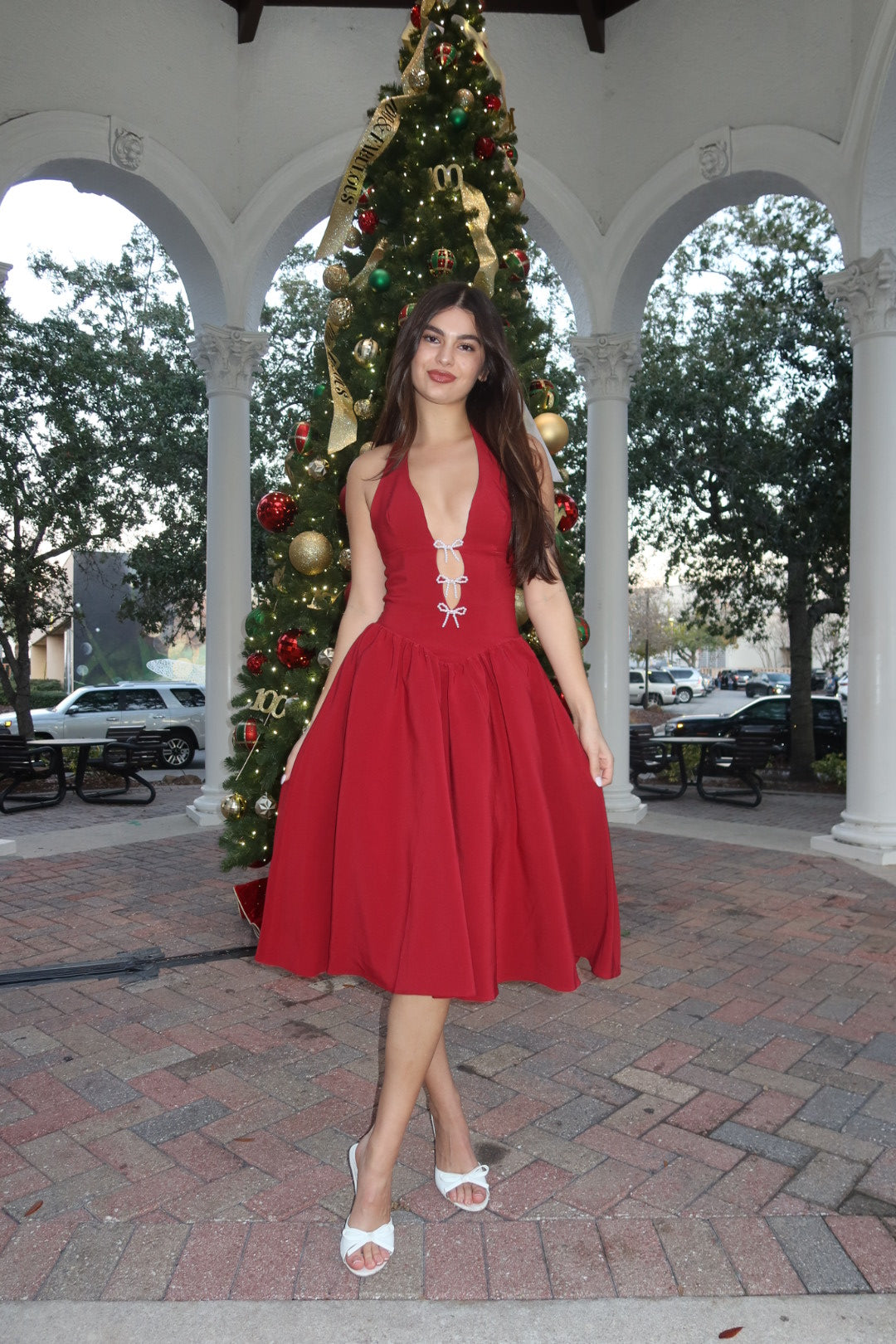 Woman in a red dress standing next to a decorated Christmas tree outdoors.